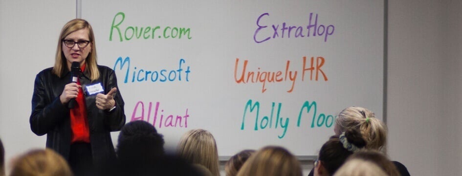 Woman giving a presentation on a stage with a microphone and a whiteboard behind her.