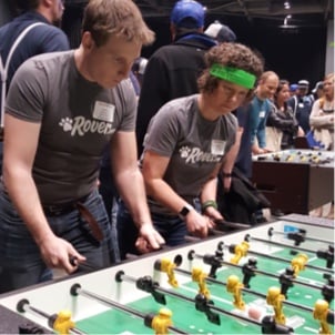 A male Rover employee and a female Rover employee intently playing a game of foosball, with another ongoing foosball game visible in the background.