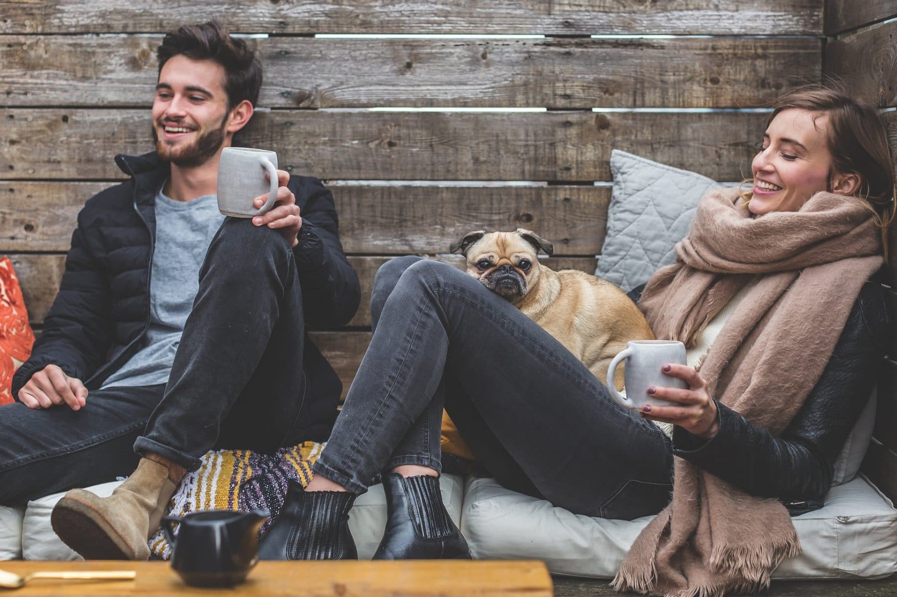 A man and woman enjoying coffee outdoors. A Pug sits on the woman's lap.