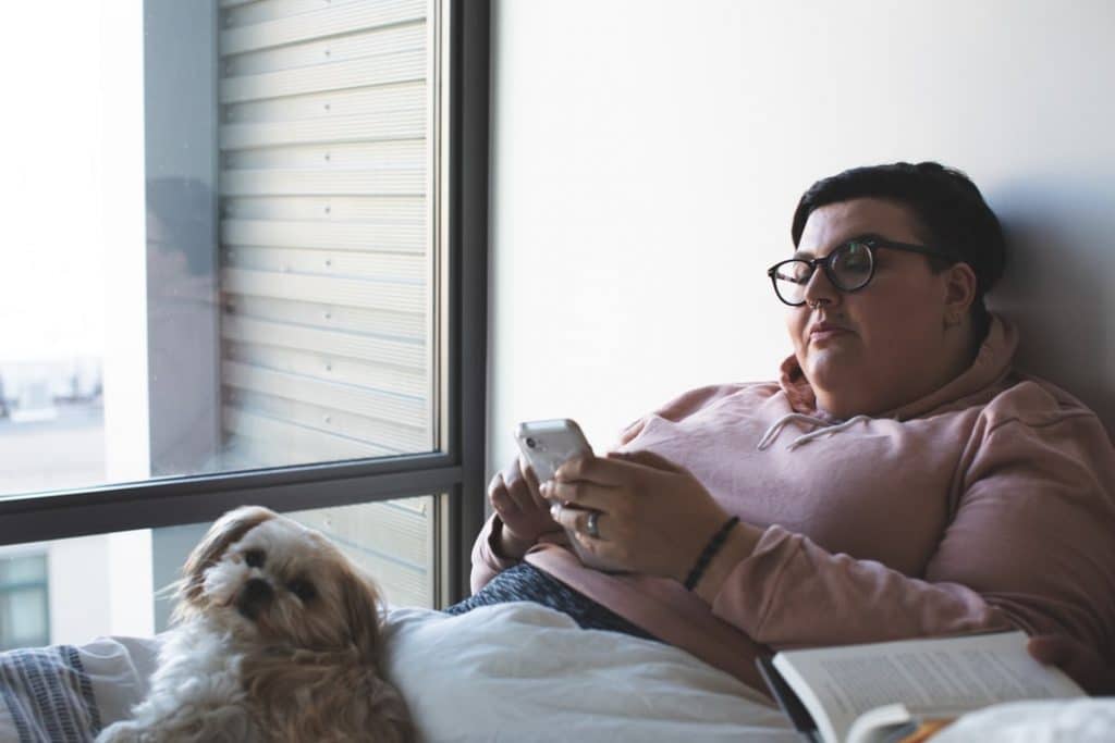 A woman looks at her phone in bed with her cozy dog