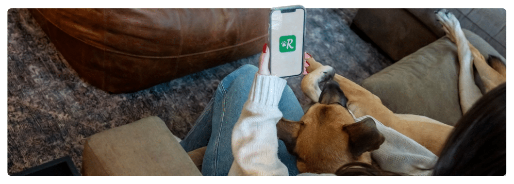 Person sitting on couch with dog opening Rover app on phone
