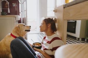 Person with bowl sits on floor with dog