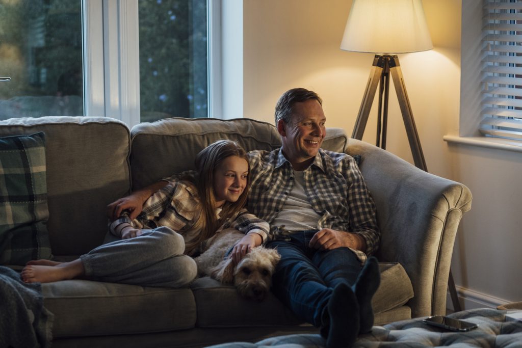 A mid-adult caucasian father lying down on the sofa with his young daughter, they are watching a movie on the tv.