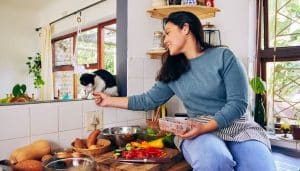 Woman sits on counter, feeding cat scraps from dinner preparation