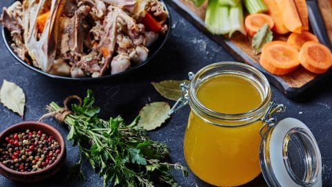 cooked chicken stock in a glass jar on a kitchen table with bouquet garni and chicken bones in a bowl, horizontal view from above