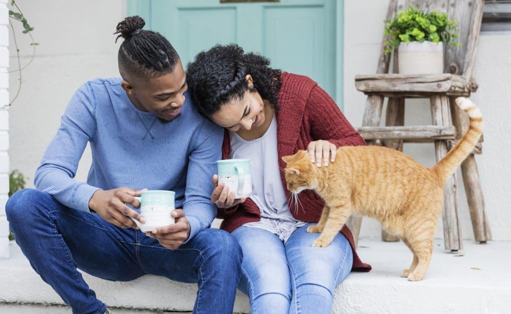 A young couple hanging out together, drinking coffee on the front steps of their home, relaxing and petting a cat