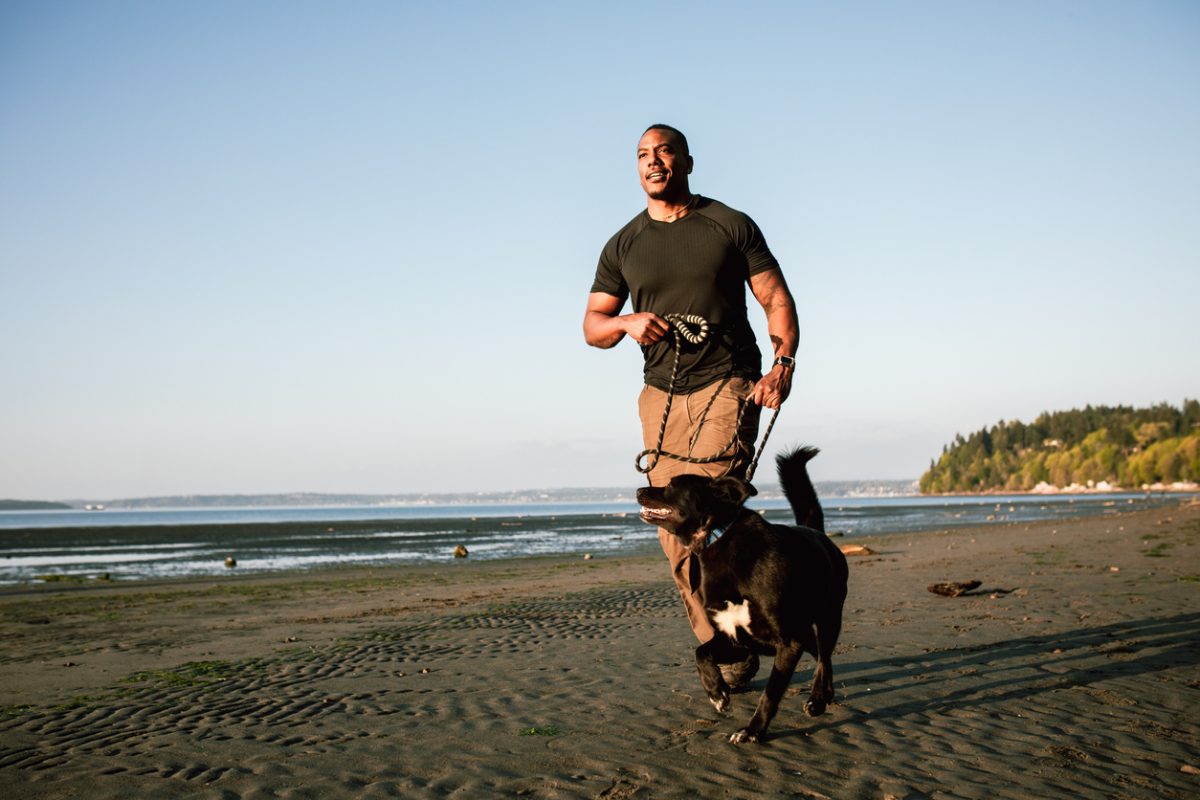 A man enjoys the Pacific Northwest, exploring a sunny Puget Sound beach in Washington state with his German Shepherd mix dog. Fun adventure and discovery with his best friend.