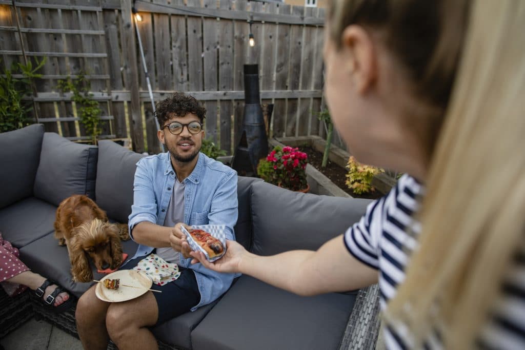 Group of friends having a bbq social gathering outdoors in the North East of England. They are sitting together sharing food on an outdoor patio and one woman is handing a man a hot-dog.