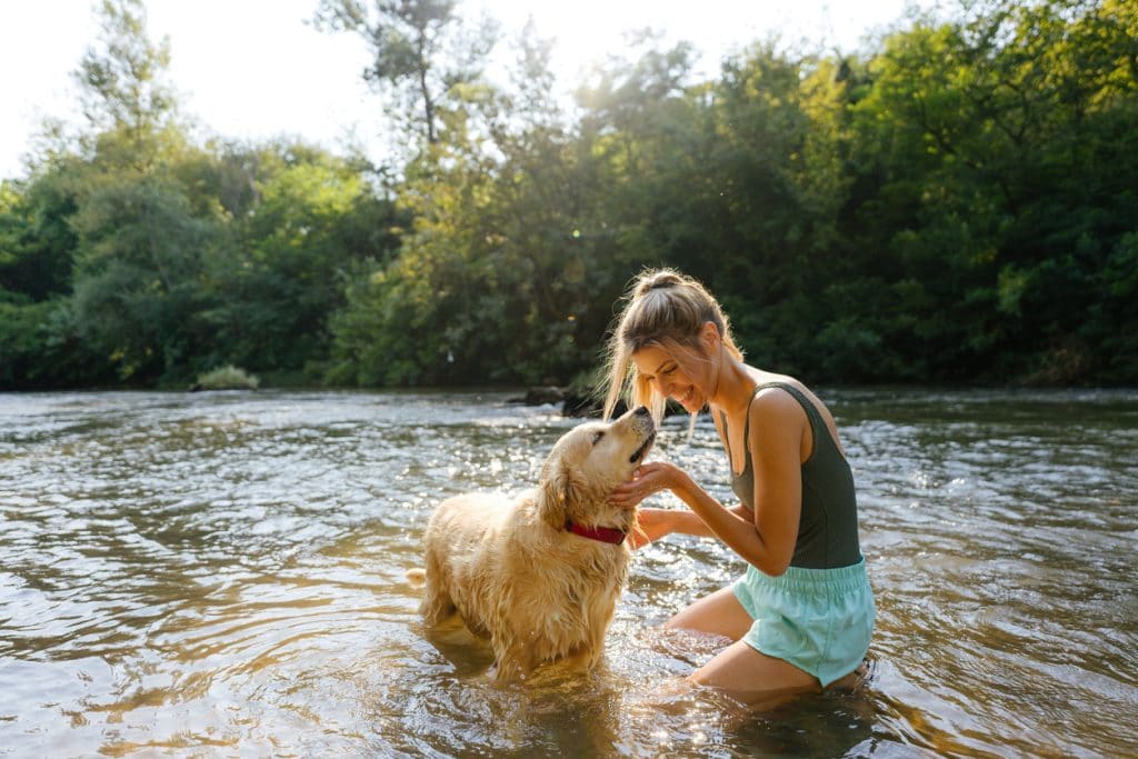 Foto de una joven sonriente y su perro bañándose en el río; pasándolo bien y disfrutando de la soleada tarde al aire libre.